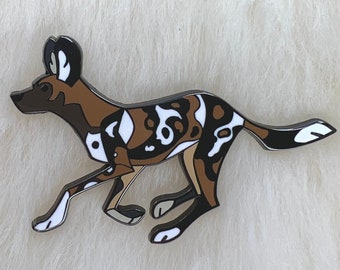 Könnte beinhalten: Eine Emaille-Anstecknadel, die einen Afrikanischen Wildhund zeigt, der rennt. Der Hund ist braun, schwarz und weiß mit einem geometrischen Muster.