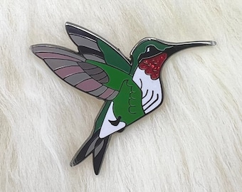Könnte beinhalten: Eine grüne und rote Emaille-Hummingbird-Anstecknadel mit weißem Bauch und schwarzen Flügeln.