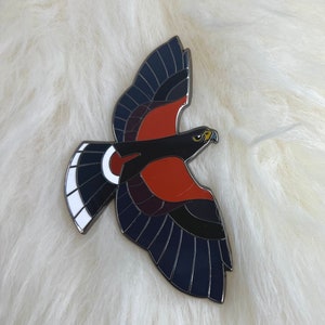 May include: A black and red enamel pin of a hawk in flight. The hawk has white markings on its tail feathers and a yellow beak.