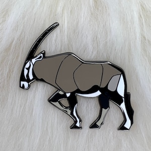Könnte beinhalten: Eine schwarze und weiße Emaille-Anstecknadel, die eine Oryx darstellt, eine Antilope mit langen, geraden Hörnern. Die Oryx steht auf einer weißen, flauschigen Oberfläche.