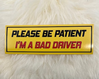 May include: Yellow and black bumper sticker with the text "PLEASE BE PATIENT I'M A BAD DRIVER" in black and red letters.