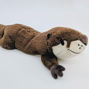 May include: A plush otter toy featuring soft brown fur on its back and tail, with a contrasting cream-colored face and underbelly. The stuffed animal has embroidered black eyes, a dark brown nose, and a stitched mouth, posed in a lying position.