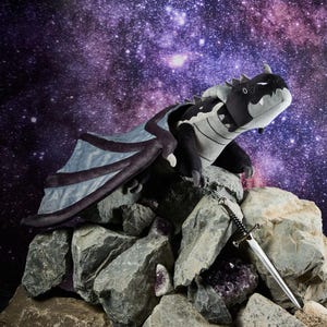 May include: A plush dragon toy with black, gray, and blue coloring, perched on a pile of rocks. The dragon has large wings and a detailed design. A silver sword rests on the rocks. The background features a starry galaxy.