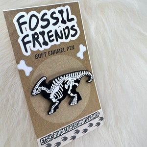 Puede incluir: Un pin de esmalte negro y blanco con un esqueleto de dinosaurio. El pin est&aacute; en una tarjeta marr&oacute;n con el texto "Fossil Friends" y "Soft Enamel Pin". La tarjeta tambi&eacute;n tiene el texto "Etsy: RoaminBisonWorkshop" y una serie de huellas de patas.