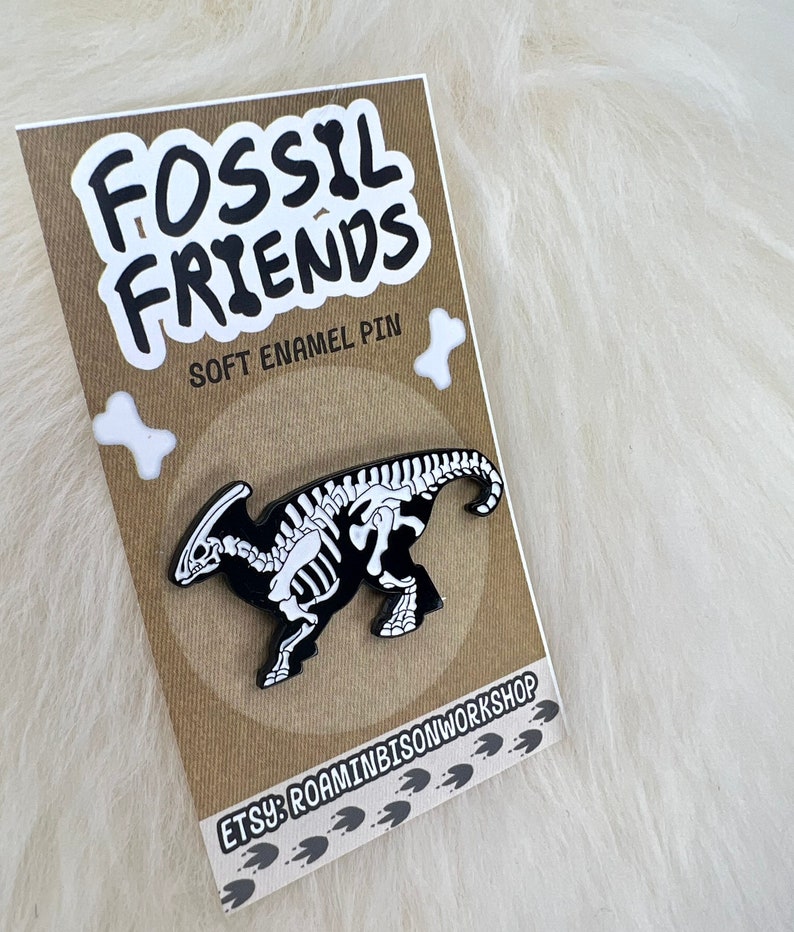 Puede incluir: Un pin de esmalte negro y blanco con un esqueleto de dinosaurio. El pin est&aacute; en una tarjeta marr&oacute;n con el texto "Fossil Friends" y "Soft Enamel Pin". La tarjeta tambi&eacute;n tiene el texto "Etsy: RoaminBisonWorkshop" y una serie de huellas de patas.