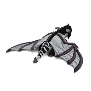 May include: A plush dragon toy in black, gray, and white. The dragon has large, outstretched wings with dark gray edges and a striped tail. The dragon's head is black with white accents, and it is lying on a white surface.