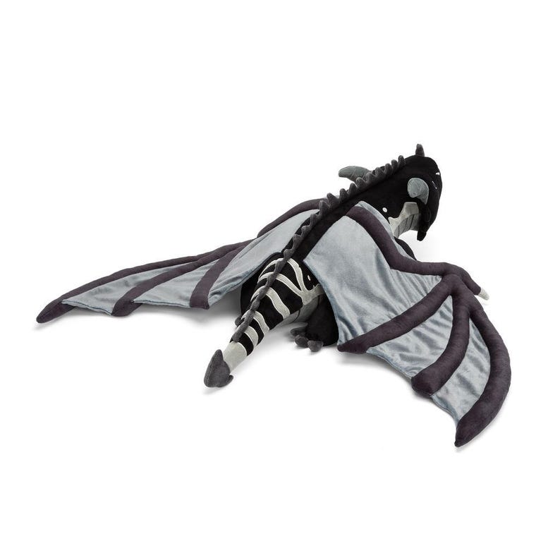 May include: A plush dragon toy in black, gray, and white. The dragon has large, outstretched wings with dark gray edges and a striped tail. The dragon's head is black with white accents, and it is lying on a white surface.