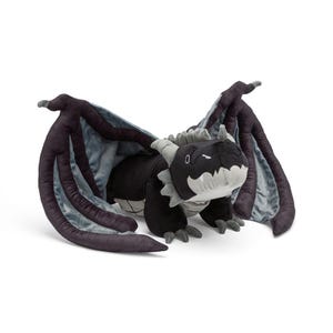 Può includere: Un giocattolo di drago di peluche in nero, grigio e azzurro. Il drago ha grandi ali, una faccia sorridente e piccole corna. Le ali sono grigio scuro con fodera azzurra. Il drago è sdraiato.