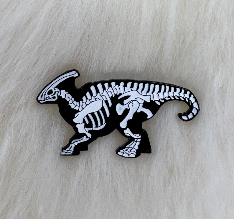 Puede incluir: Pin de esmalte negro y blanco de un esqueleto de dinosaurio. El dinosaurio est&aacute; en una pose de carrera con la cabeza girada hacia un lado.