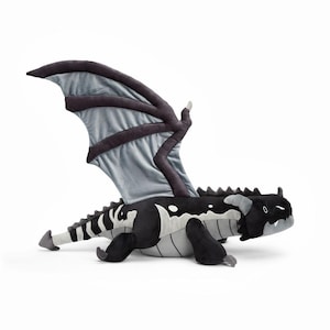May include: A plush dragon toy in black, gray, and white. The dragon has large wings, a long tail, and a detailed design. The dragon is lying down on a white surface.
