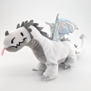 Könnte beinhalten: Ein weißes Plüschdrachenspielzeug mit grauen Akzenten, darunter ein stacheliger Rücken und ein lächelndes Gesicht mit einer rosa Zunge. Der Drache hat schillernde silberne Flügel und steht auf allen Vieren.