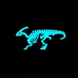 Puede incluir: Una figurita de esqueleto de dinosaurio azul que brilla en la oscuridad. El dinosaurio est&aacute; de pie sobre sus cuatro patas con la cola enroscada.