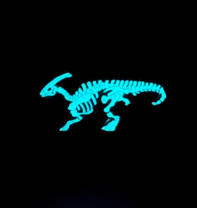 Puede incluir: Una figurita de esqueleto de dinosaurio azul que brilla en la oscuridad. El dinosaurio est&aacute; de pie sobre sus cuatro patas con la cola enroscada.