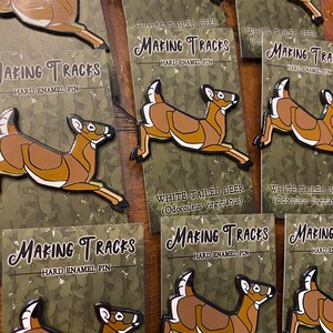 May include: A set of enamel pins featuring a white-tailed deer in various poses. The pins are brown and white with black outlines and have the text "Making Tracks" and "Hard Enamel Pin" on the cards they are displayed on.