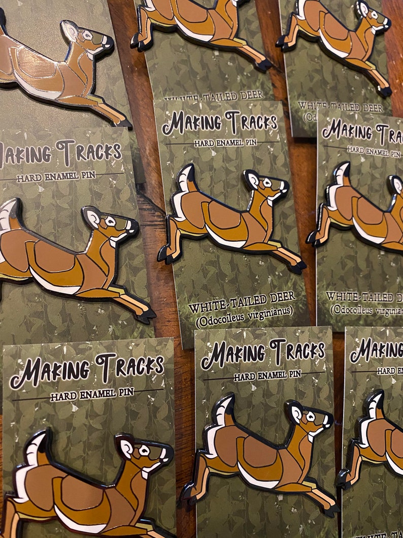 May include: A set of enamel pins featuring a white-tailed deer in various poses. The pins are brown and white with black outlines and have the text "Making Tracks" and "Hard Enamel Pin" on the cards they are displayed on.