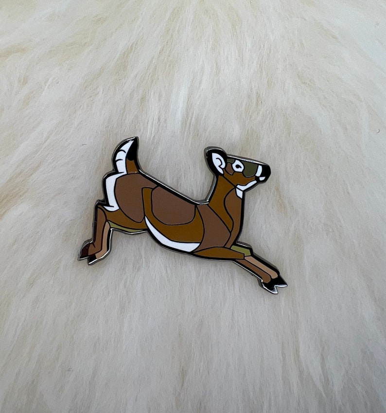 May include: A brown and white enamel pin of a stylized deer leaping in mid-air.