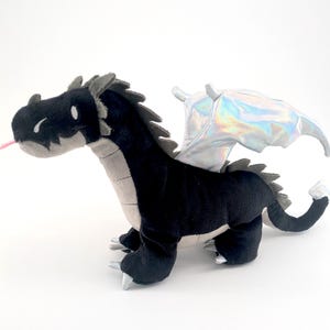 May include: A plush dragon toy in black and gray with iridescent wings. The dragon has a pink tongue sticking out and silver claws. The toy is made of soft fabric and is designed to look like a friendly dragon.