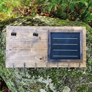 May include: A rustic wooden key holder with a chalkboard insert and hooks. The chalkboard has four white lines drawn across it. The key holder has a dark brown stain and is made of reclaimed wood.