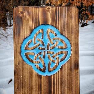 May include: A wooden sign with a blue Celtic knot design carved into the surface. The wood has a dark brown stain.