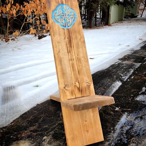 May include: A wooden chair with a Celtic knot design carved into the back. The chair is made of natural wood and has a rustic finish. The chair is sitting on a snowy ground.