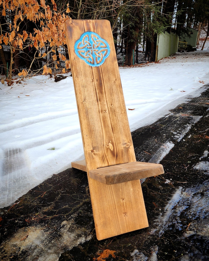 May include: A wooden chair with a Celtic knot design carved into the back. The chair is made of natural wood and has a rustic finish. The chair is sitting on a snowy ground.