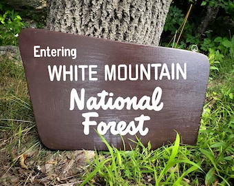 May include: A trapezoidal brown wooden sign with white lettering that reads Entering WHITE MOUNTAIN National Forest. The sign features a smooth painted finish and a small metal hanging hook attached to the top right corner of the board.