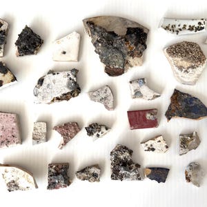 May include: A collection of various ceramic shards and pottery fragments in assorted shapes and sizes. The pieces feature diverse textures, including smooth glazed surfaces in white, pink, red, and blue, alongside rough, weathered, and dark edges.
