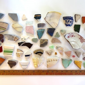May include: A collection of ceramic shards in various shapes and colors, including blue, white, orange, and green. Some pieces have decorative patterns. A wooden ruler with the text "A Barrel of Satisfaction in Every Gallon" is at the bottom.