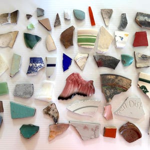 May include: A collection of ceramic shards in various shapes and colors, including white, blue, green, red, and brown. Some pieces have patterns or designs, and one shard has the text "well Dish".