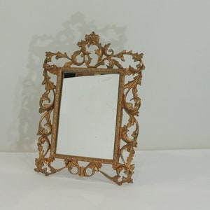 May include: An ornate metal frame in a gold tone, featuring a small rectangular mirror. The frame has a decorative design with intricate details.