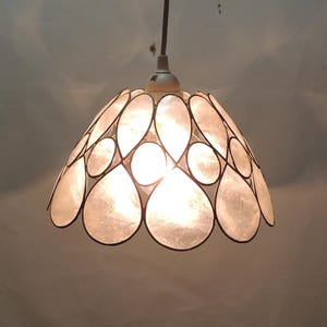 May include: A pendant light fixture with a white, petal-shaped lampshade. The shade is outlined in black and has a warm, glowing light emanating from within. The fixture hangs from a white ceiling mount.