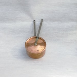 Puede incluir: Una olla pequeña de cobre con tapa y dos asas metálicas. La olla está sobre una superficie blanca.