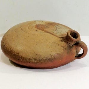 Könnte beinhalten: Eine runde Steingutflasche mit kleinem Ausguss und Griff. Die Flasche hat einen hellbraunen Körper mit dunkelbraunen und rotbraunen Akzenten. Die Oberseite weist ein Spiralmuster auf. Die Flasche ist wahrscheinlich ein antikes oder Vintage-Objekt.