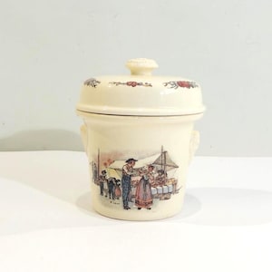 Puede incluir: Tarro de cerámica color crema con tapa, con una ilustración detallada de una escena de mercado. El tarro tiene una pequeña asa a cada lado y detalles florales en la tapa. La ilustración representa a personas en un puesto de mercado.