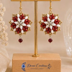 May include: A pair of star-shaped beaded earrings featuring alternating white and deep red petal-shaped beads, a central clear crystal, and gold-toned accents. Each earring hangs from a gold-toned hook and includes a small red crystal drop.