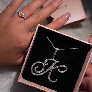 Puede incluir: Un collar con inicial de diamantes en una caja negra con interior rosa. El collar tiene la forma de la letra "K" y está incrustado con pequeños diamantes. Un anillo de diamantes está en un dedo en el fondo.