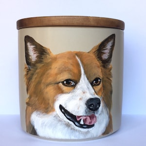 Puede incluir: Un tarro de cerámica con tapa de madera, pintado a mano con un retrato de un perro. El perro tiene pelo marrón y blanco, con un hocico blanco y una lengua rosa que sobresale. El tarro es de color beige claro.