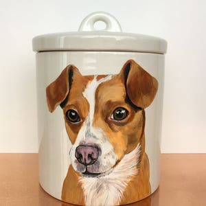 Op de afbeelding: Witte keramische pot met een deksel en een handgeschilderd portret van een bruine en witte hond met bruine ogen. De hond kijkt recht naar de kijker.
