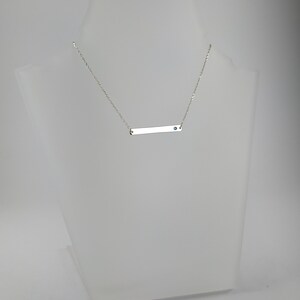 Könnte beinhalten: Eine silberne Halskette mit einem dünnen Barrenanhänger und einem kleinen blauen Edelstein. Die Halskette befindet sich auf einer weißen Puppe.