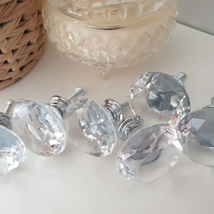 May include: Six clear crystal cabinet knobs with silver metal bases. The knobs have a faceted design and are arranged on a white surface.
