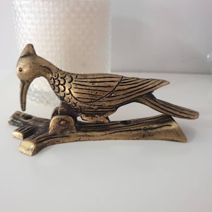 May include: A gold-toned metal door knocker shaped like a bird perched on a branch. The bird has detailed wings and a beak.