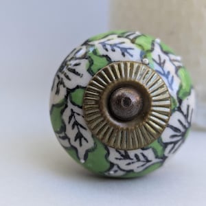May include: Green ceramic knob with a white floral pattern and a brass centre. The knob has a raised, textured design.