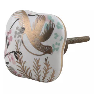 May include: White ceramic cabinet knob with a gold bird and floral design. The knob has a square shape with rounded edges and a gold screw.