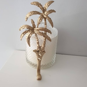 May include: Gold palm tree shaped wall hook. The hook is in the shape of two palm trees with a single stem. The hook is attached to a white wall.