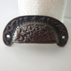 Peut inclure: Une poignée de tiroir en fonte de couleur brun foncé avec un motif floral. La poignée a une forme incurvée et deux trous pour le montage.