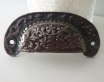 May include: A dark brown cast iron drawer pull with a floral design. The pull has a curved shape and two holes for mounting.