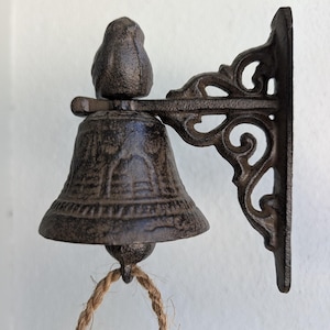 Könnte beinhalten: Eine Glocke aus Gusseisen mit rustikalem Finish, montiert an einer dekorativen Halterung. Die Glocke hat einen Seilzug und ist für die Wandmontage konzipiert. Die Abmessungen der Glocke betragen ca. 10 cm Höhe und 7,6 cm Breite.