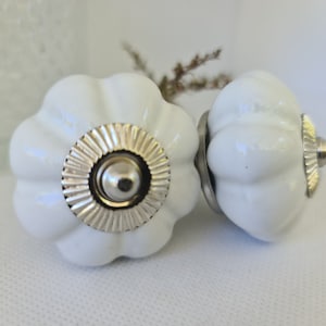 Peut inclure: Deux boutons de tiroir en céramique blanche en forme de citrouille avec des accents argentés. Les boutons ont une surface texturée en relief et un anneau argenté autour du centre.