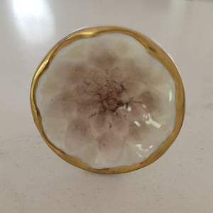 Op de afbeelding: Een witte keramische knop met een gouden rand en een reliëf bloemmotief. De knop heeft een subtiele kleurvariatie in bruin en wit.