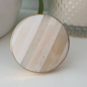 May include: A round, white and beige striped knob with a gold metal rim. The knob is a decorative hardware piece.